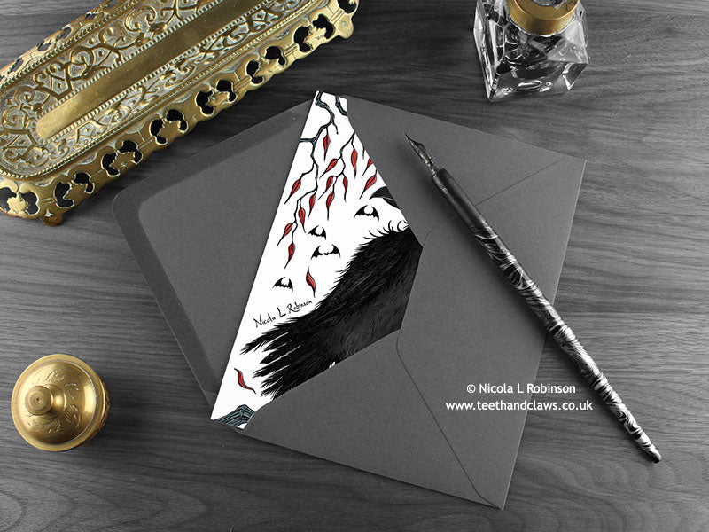 Gothic crow greeting card, shown lying flat, with my original pen and ink illustration of a crow, inside an open dark grey envelope. With vintage Victorian pen and writing desk accessories. Design and illustration © Nicola L Robinson | Teeth and Claws