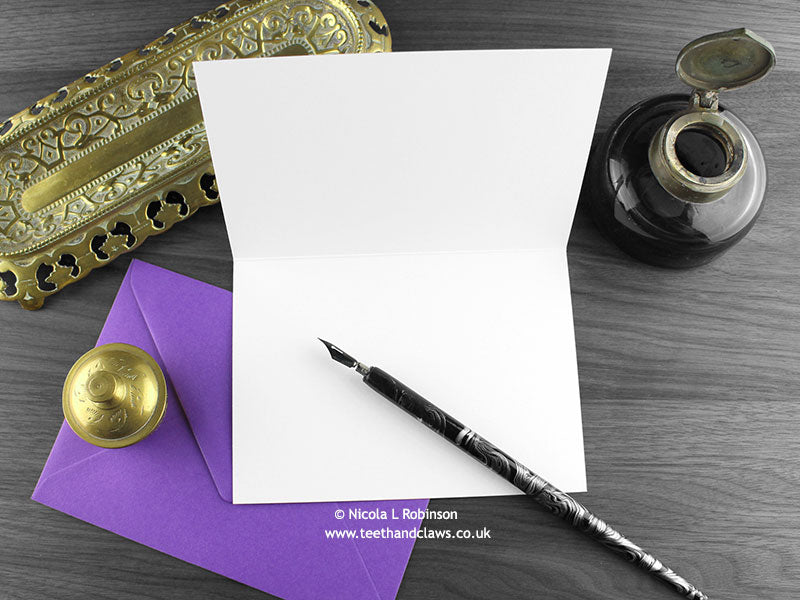 Crow greeting card, shown open and flat with blank inside, on a purple envelope, alongside vintage Victorian pen and writing desk accessories. Design and illustration © Nicola L Robinson | Teeth and Claws