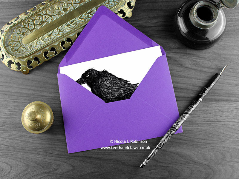 Crow greeting card, shown lying flat, with my original pen and ink illustration of a crow, inside an open purple envelope. With vintage Victorian pen and writing desk accessories. Design and illustration © Nicola L Robinson | Teeth and Claws