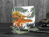 Stegosaurus and ferns greeting card, front view, featuring my original pen and ink dinosaur illustration. The card is standing on a green envelope, shown at an angle. Design and illustration © Nicola L Robinson | Teeth and Claws