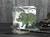 Triceratops and ferns greeting card, front view, featuring my original pen and ink dinosaur illustration. The card is standing on a green envelope, shown at an angle. Design and illustration © Nicola L Robinson | Teeth and Claws