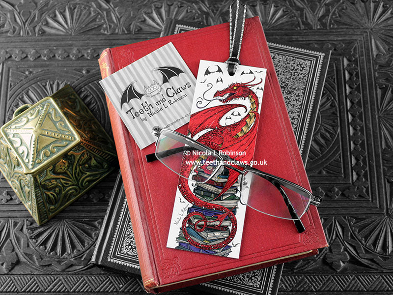 Dragon bookmark featuring my original pen and ink illustration, front view of the bookmark resting on books with one of my branded cards. Made in the UK. Design and illustration © Nicola L Robinson | Teeth and Claws.