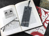 Dragon bookmark featuring my original pen and ink illustration, back view of the bookmark resting on an open book with reading glasses and one of my branded cards. Made in the UK. Design and illustration © Nicola L Robinson | Teeth and Claws.
