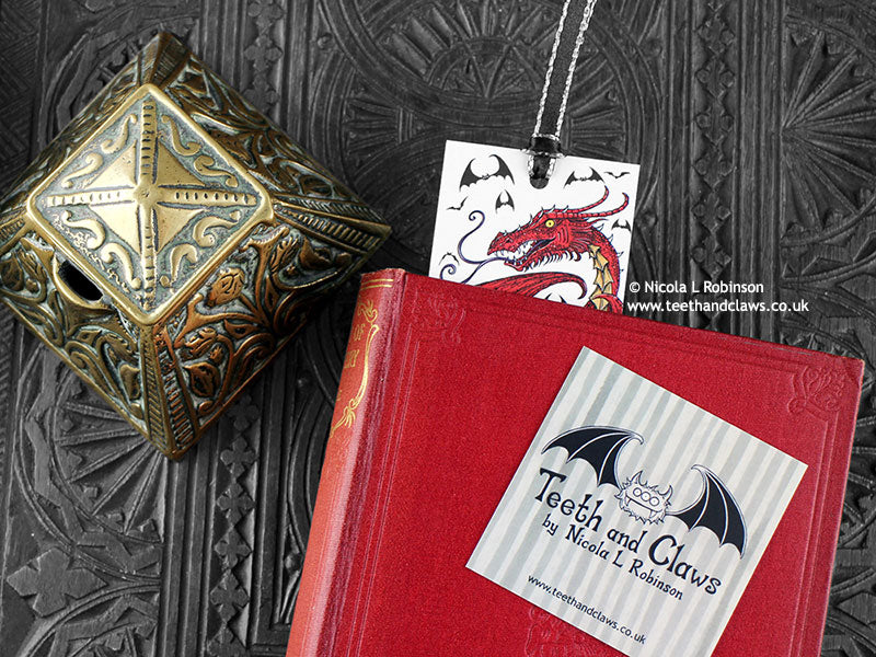 Dragon bookmark featuring my original pen and ink illustration, front view with the top showing from inside a closed book with one of my branded cards. Made in the UK. Design and illustration © Nicola L Robinson | Teeth and Claws.