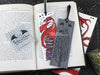 Dragon bookmark featuring my original pen and ink illustrations, front and back view of the bookmark resting on an open book with one of my branded cards. Made in the UK. Design and illustration © Nicola L Robinson | Teeth and Claws.