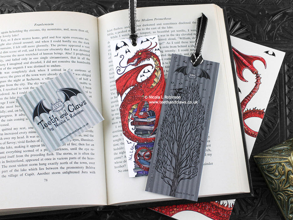 Dragon bookmark featuring my original pen and ink illustrations, front and back view of the bookmark resting on an open book with one of my branded cards. Made in the UK. Design and illustration © Nicola L Robinson | Teeth and Claws.