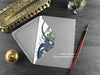Dragon Dad Greeting Card, shown lying flat, with my original pen and ink illustration of a blue dragon and little green dragon, inside an open grey envelope. With vintage Victorian pen and writing desk accessories. Design and illustration © Nicola L Robinson | Teeth and Claws