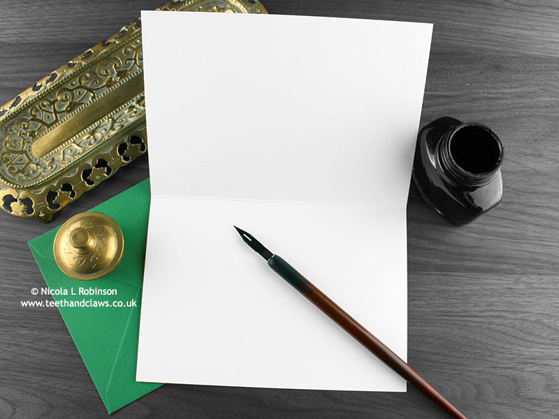 Deinonychus greeting card, shown open and flat with blank inside, on a green envelope, alongside vintage Victorian pen and writing desk accessories. Design and illustration © Nicola L Robinson | Teeth and Claws
