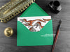 Dinosaur greeting card, shown lying flat, with my original pen and ink illustration of Deinonychus, inside an open green envelope. With vintage Victorian pen and writing desk accessories. Design and illustration © Nicola L Robinson | Teeth and Claws