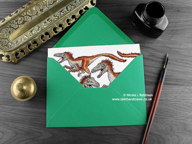 Dinosaur greeting card, shown lying flat, with my original pen and ink illustration of Deinonychus, inside an open green envelope. With vintage Victorian pen and writing desk accessories. Design and illustration © Nicola L Robinson | Teeth and Claws