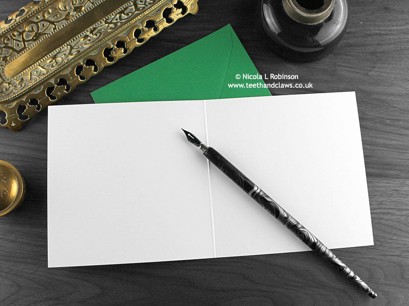 Dinosaur Greeting card, shown open and flat with blank inside, on a green envelope, alongside vintage Victorian pen and writing desk accessories. Design and illustration © Nicola L Robinson | Teeth and Claws