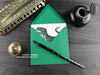 Dinosaur greeting card, shown lying flat, with my original pen and ink illustration of a big T rex and a baby T rex, inside an open green envelope. With vintage Victorian pen and writing desk accessories. Design and illustration © Nicola L Robinson | Teeth and Claws