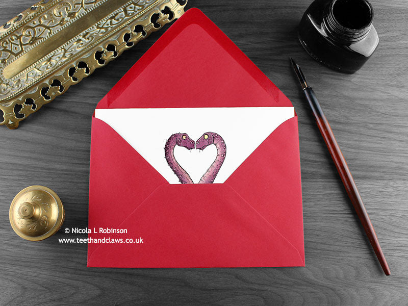 Dinosaur love card, shown lying flat, with my original pen and ink illustration of two dinosaurs, inside an open red envelope. With vintage Victorian pen and writing desk accessories. Design and illustration © Nicola L Robinson | Teeth and Claws