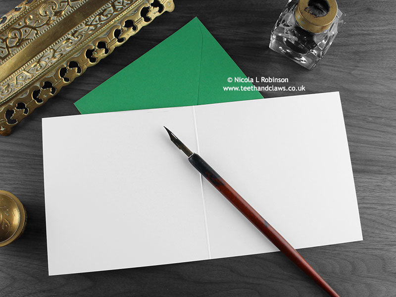 Dinosaur Birthday card, shown open and flat with blank inside, on a green envelope, alongside vintage Victorian pen and writing desk accessories. Design and illustration © Nicola L Robinson | Teeth and Claws