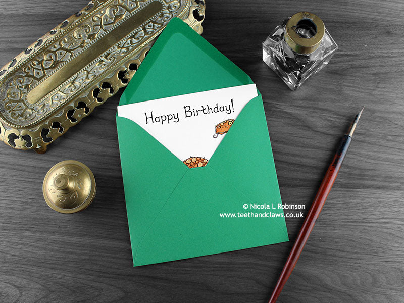 Dinosaur Birthday card, shown lying flat, with my original pen and ink illustration of a diplodocus, inside an open green envelope. With vintage Victorian pen and writing desk accessories. Design and illustration © Nicola L Robinson | Teeth and Claws