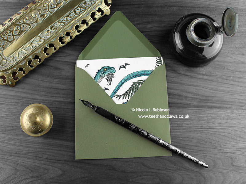 Diplodocus and ferns greeting card, shown lying flat, with my original pen and ink illustration of a dinosaur, inside an open green envelope. With vintage Victorian pen and writing desk accessories. Design and illustration © Nicola L Robinson | Teeth and Claws