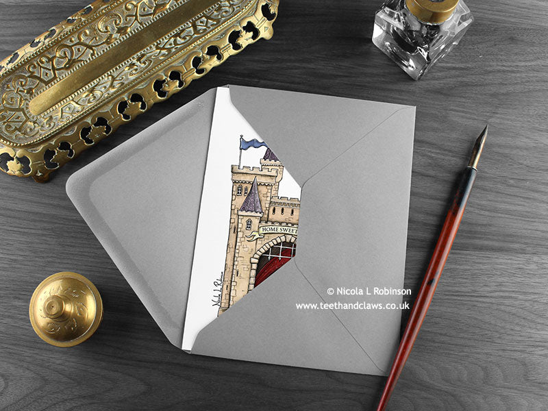 Dragon Castle New Home card, shown lying flat, with my original pen and ink illustration of a castle with Welsh dragon, inside an open grey envelope. With vintage Victorian pen and writing desk accessories. Design and illustration © Nicola L Robinson | Teeth and Claws