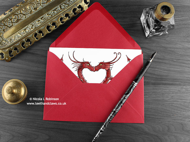 Dragon love greeting card, shown lying flat, with my original pen and ink illustration of two red dragons, inside an open red envelope. With vintage Victorian pen and writing desk accessories. Design and illustration © Nicola L Robinson | Teeth and Claws