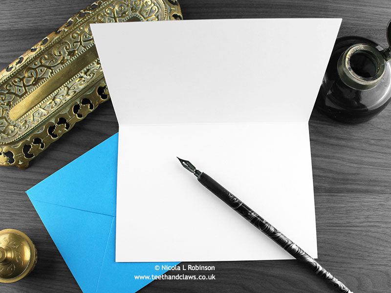 Gothic fish greeting card, shown open and flat with blank inside, on a blue envelope, alongside vintage Victorian pen and writing desk accessories. Design and illustration © Nicola L Robinson | Teeth and Claws
