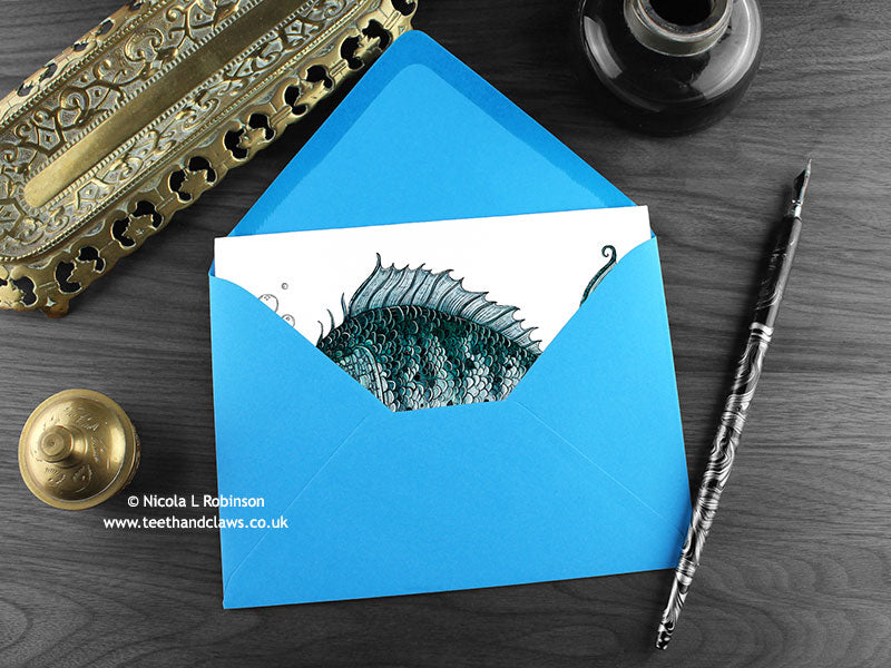 Fish greeting card, shown lying flat, with my original pen and ink illustration of a Gothic fish, inside an open blue envelope. With vintage Victorian pen and writing desk accessories. Design and illustration © Nicola L Robinson | Teeth and Claws