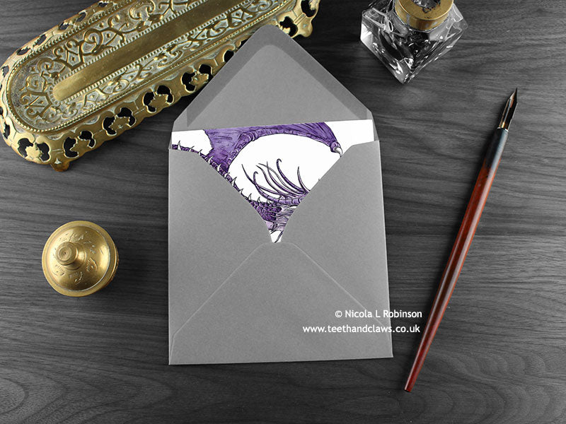 Flying Dragons Greeting Card, shown lying flat, with my original pen and ink illustration of flying dragons, inside an open grey envelope. With vintage Victorian pen and writing desk accessories. Design and illustration © Nicola L Robinson | Teeth and Claws
