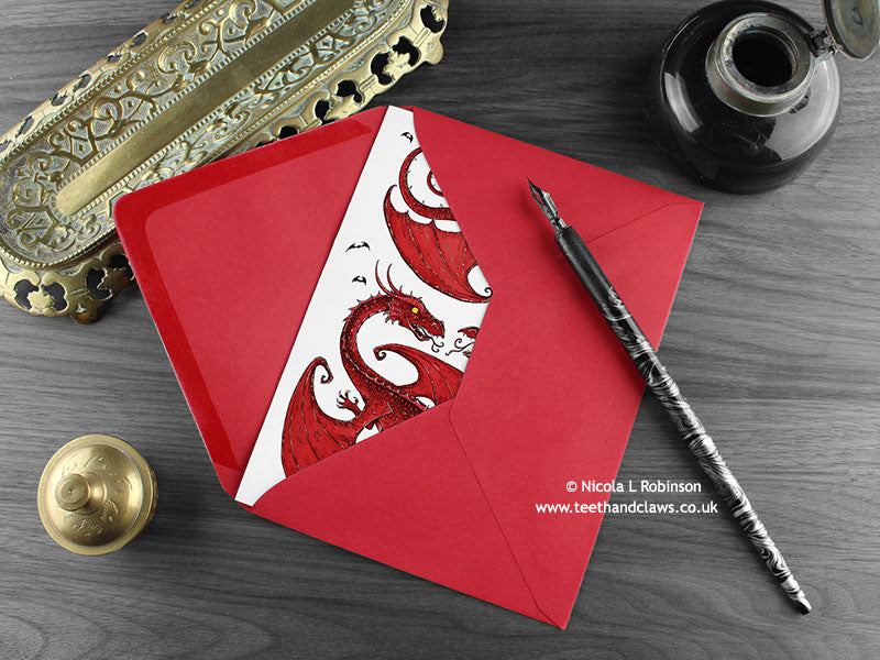 Two Dragons love card, shown lying flat, with my original pen and ink illustration of two red dragons, inside an open red envelope. With vintage Victorian pen and writing desk accessories. Design and illustration © Nicola L Robinson | Teeth and Claws