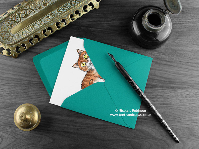 Ginger and white tabby cat greeting card, shown lying flat, with my original pen and ink illustration of a ginger and white cat, inside an open teal envelope. With vintage Victorian pen and writing desk accessories. Design and illustration © Nicola L Robinson | Teeth and Claws