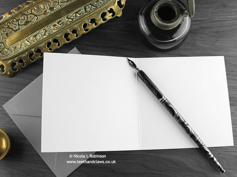 Green Dragon with Dragon Eggs Greeting Card, shown open and flat with blank inside, on a grey envelope, alongside vintage Victorian pen and writing desk accessories. Design and illustration © Nicola L Robinson | Teeth and Claws