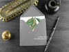 Green Dragon with Dragon Eggs Greeting Card, shown lying flat, with my original pen and ink illustration of a green dragon, inside an open grey envelope. With vintage Victorian pen and writing desk accessories. Design and illustration © Nicola L Robinson | Teeth and Claws