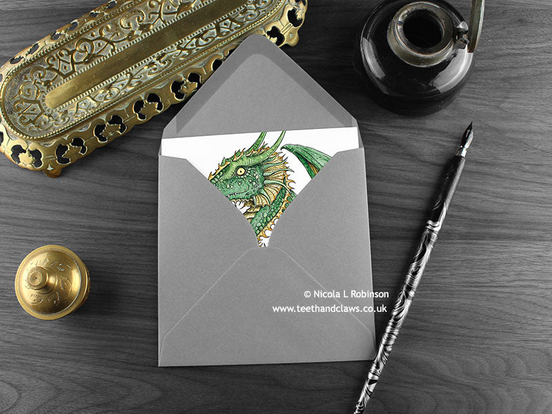 Green Dragon with Dragon Eggs Greeting Card, shown lying flat, with my original pen and ink illustration of a green dragon, inside an open grey envelope. With vintage Victorian pen and writing desk accessories. Design and illustration © Nicola L Robinson | Teeth and Claws