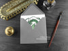 Green Horned Dragon Greeting Card, shown lying flat, with my original pen and ink illustration of a green dragon, inside an open grey envelope. With vintage Victorian pen and writing desk accessories. Design and illustration © Nicola L Robinson | Teeth and Claws
