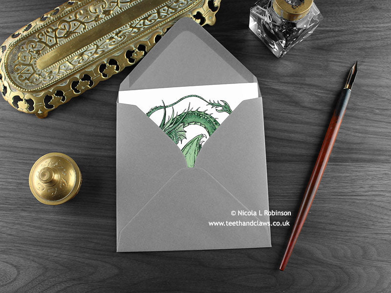 Green Horned Dragon Greeting Card, shown lying flat, with my original pen and ink illustration of a green dragon, inside an open grey envelope. With vintage Victorian pen and writing desk accessories. Design and illustration © Nicola L Robinson | Teeth and Claws