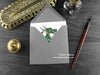 Green Dragon Greeting Card, shown lying flat, with my original pen and ink illustration of a green dragon, inside an open grey envelope. With vintage Victorian pen and writing desk accessories. Design and illustration © Nicola L Robinson | Teeth and Claws