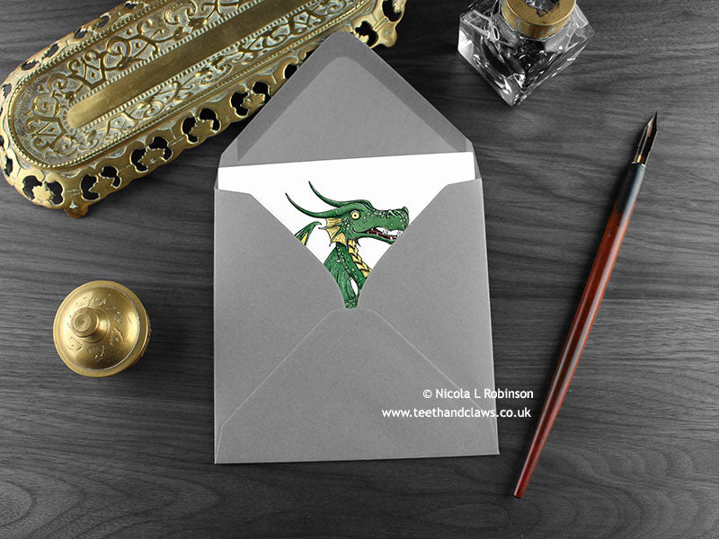 Green Dragon Greeting Card, shown lying flat, with my original pen and ink illustration of a green dragon, inside an open grey envelope. With vintage Victorian pen and writing desk accessories. Design and illustration © Nicola L Robinson | Teeth and Claws