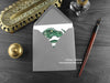 Green Celtic Spiral Dragon Greeting Card, shown lying flat, with my original pen and ink illustration of a green dragon, inside an open grey envelope. With vintage Victorian pen and writing desk accessories. Design and illustration © Nicola L Robinson | Teeth and Claws