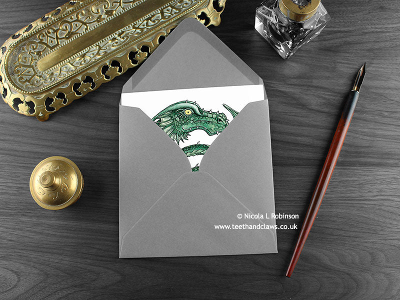 Green Celtic Spiral Dragon Greeting Card, shown lying flat, with my original pen and ink illustration of a green dragon, inside an open grey envelope. With vintage Victorian pen and writing desk accessories. Design and illustration © Nicola L Robinson | Teeth and Claws