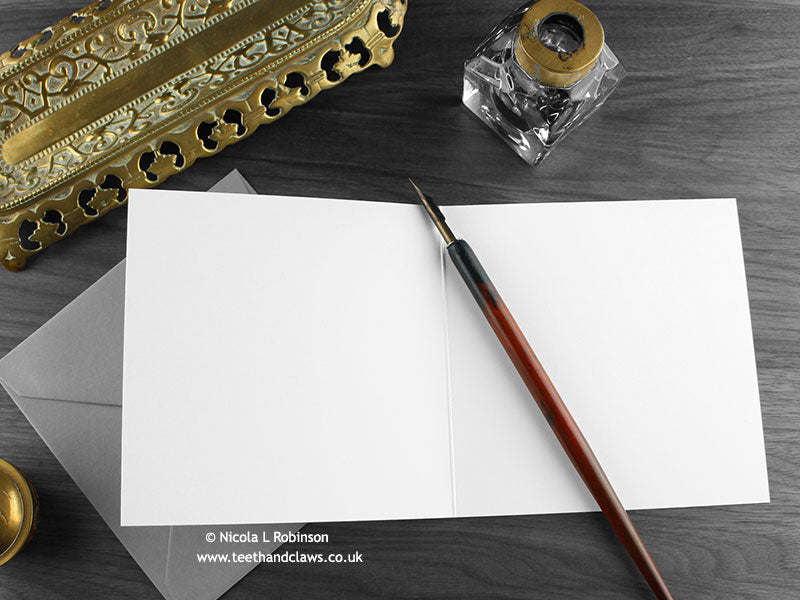 Dark Inky Dragon Greeting Card, shown open and flat with blank inside, on a grey envelope, alongside vintage Victorian pen and writing desk accessories. Design and illustration © Nicola L Robinson | Teeth and Claws