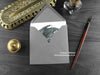 Dark Inky Dragon Greeting Card, shown lying flat, with my original pen and ink illustration of a dark grey dragon, inside an open grey envelope. With vintage Victorian pen and writing desk accessories. Design and illustration © Nicola L Robinson | Teeth and Claws