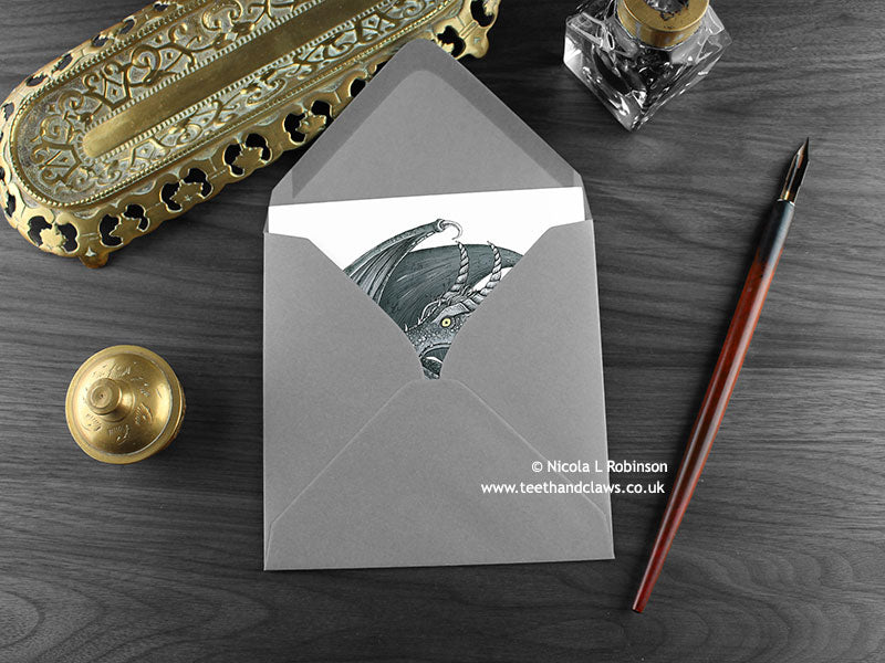 Dark Inky Dragon Greeting Card, shown lying flat, with my original pen and ink illustration of a dark grey dragon, inside an open grey envelope. With vintage Victorian pen and writing desk accessories. Design and illustration © Nicola L Robinson | Teeth and Claws