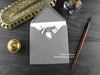 Lilac Dragon Greeting Card, shown lying flat, with my original pen and ink illustration of a purple dragon, inside an open grey envelope. With vintage Victorian pen and writing desk accessories. Design and illustration © Nicola L Robinson | Teeth and Claws