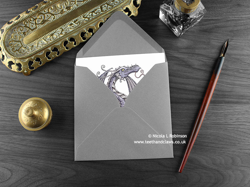 Lilac Dragon Greeting Card, shown lying flat, with my original pen and ink illustration of a purple dragon, inside an open grey envelope. With vintage Victorian pen and writing desk accessories. Design and illustration © Nicola L Robinson | Teeth and Claws