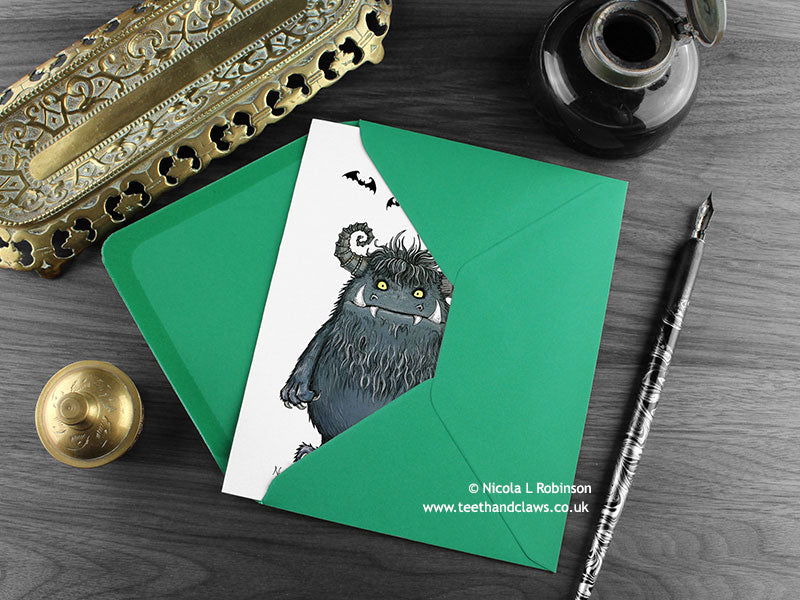 Monster greeting card, shown lying flat, with my original pen and ink illustration of a big hairy monster, inside an open green envelope. With vintage Victorian pen and writing desk accessories. Design and illustration © Nicola L Robinson | Teeth and Claws