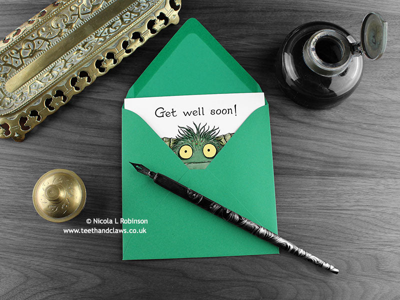 Monster Get Well Soon card, shown lying flat, with my original pen and ink illustration of a hairy monster, inside an open green envelope. With vintage Victorian pen and writing desk accessories. Design and illustration © Nicola L Robinson | Teeth and Claws