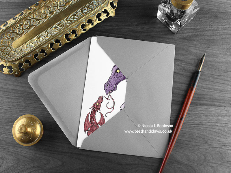 Dragon Mum Greeting Card, shown lying flat, with my original pen and ink illustration of a little red dragon and purple dragon, inside an open grey envelope. With vintage Victorian pen and writing desk accessories. Design and illustration © Nicola L Robinson | Teeth and Claws