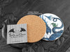 Blue Dragon Coaster featuring my original pen and ink illustration of a blue dragon, back view showing the cork backing, alongside one of my branded cards. Made in the UK. Design and illustration © Nicola L Robinson | Teeth and Claws.