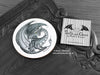 Dark Grey Dragon Coaster featuring my original pen and ink illustration of a dark grey dragon, front view alongside one of my branded cards. Made in the UK. Design and illustration © Nicola L Robinson | Teeth and Claws.