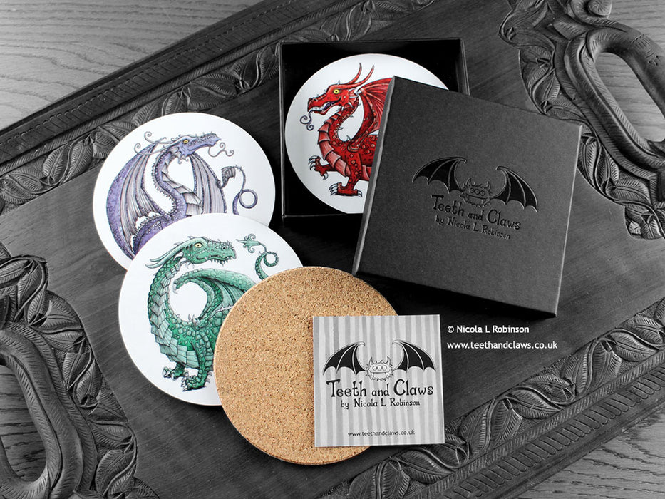 Set of four of my dragon coasters featuring my original pen and ink dragon illustrations, front view alongside a black gift box and card, with one coaster turned over to show the cork backing. Made in the UK. Design and illustration © Nicola L Robinson | Teeth and Claws.