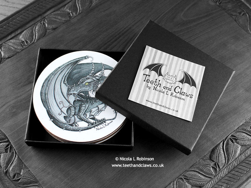 Set of four of my dragon coasters featuring my original pen and ink dragon illustrations, front view in an open black gift box with card. Made in the UK. Design and illustration © Nicola L Robinson | Teeth and Claws.
