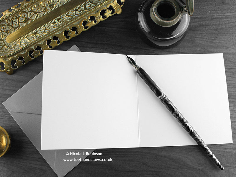 Green Friendly Dragon Greeting Card, shown open and flat with blank inside, on a grey envelope, alongside vintage Victorian pen and writing desk accessories. Design and illustration © Nicola L Robinson | Teeth and Claws