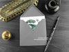 Green Friendly Dragon Greeting Card, shown lying flat, with my original pen and ink illustration of a light green dragon, inside an open grey envelope. With vintage Victorian pen and writing desk accessories. Design and illustration © Nicola L Robinson | Teeth and Claws